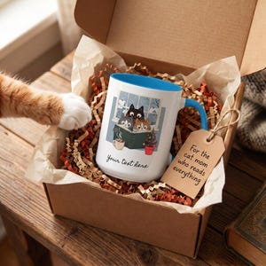 May include: A white ceramic mug with a blue interior and handle, featuring a graphic of three cats reading a book by a window, a small Christmas tree, and a mug of cocoa. The text "Your text here" is printed below, with a gift tag reading "For the cat mom who reads everything."