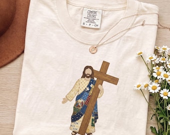 Könnte beinhalten: Ein cremefarbenes Kurzarm-T-Shirt mit einer Grafik einer Figur, die ein großes Holzkreuz hält. Die Figur trägt ein Gewand mit blauen, gelben und weißen gemusterten Abschnitten. Ein Etikett zeigt die Aufschrift COMFORT COLORS S P CH.
