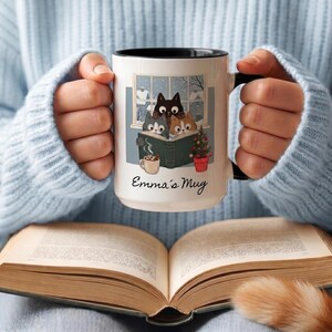 May include: A white ceramic mug with a black interior and handle, featuring a graphic of three cats reading a book in front of a window. The illustration includes a small potted Christmas tree and a mug of cocoa. The text Emma's Mug is printed below.