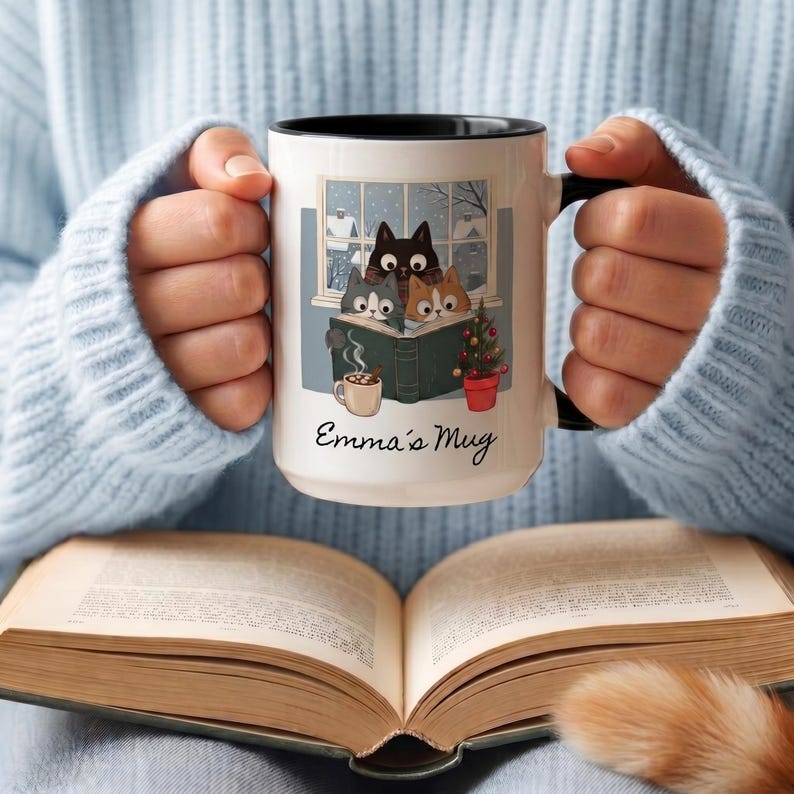 May include: A white ceramic mug with a black interior and handle, featuring a graphic of three cats reading a book in front of a window. The illustration includes a small potted Christmas tree and a mug of cocoa. The text Emma's Mug is printed below.