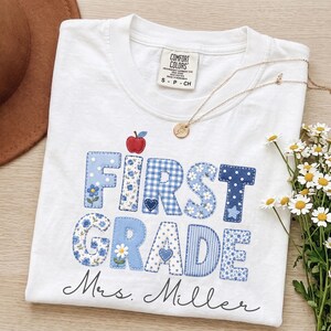 May include: A white t-shirt featuring the text FIRST GRADE in blue patterned fabric letters with a small red apple icon above. Below the text, the name Mrs. Miller is printed in black script. The shirt has a Comfort Colors tag and a gold necklace.