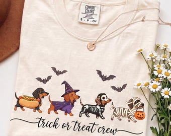 Könnte beinhalten: Ein elfenbeinfarbenes T-Shirt mit einer Grafik von vier Dackeln in Halloween-Kostümen: ein Hotdog, ein Zauberer, ein Skelett und eine Mumie mit Kürbiseimer. Über den Hunden sind vier Fledermäuse und darunter der Text "trick or treat crew" zu sehen.