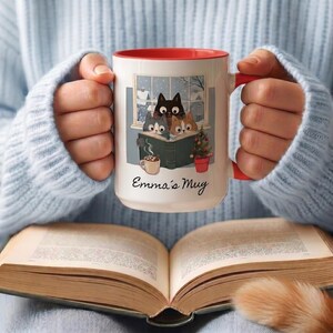 May include: A white ceramic mug with a red interior and handle, featuring a graphic of three cats reading a book by a window, a small potted Christmas tree, and a mug of cocoa. The text "Emma's Mug" is printed in black script below the illustration.