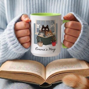 May include: A white ceramic mug with a lime green interior and handle, featuring a graphic of three cats reading a book by a window, a small mug of cocoa, and a potted plant. The text Emma's Mug is printed in black script below the illustration.