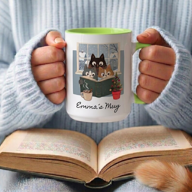 May include: A white ceramic mug with a lime green interior and handle, featuring a graphic of three cats reading a book by a window, a small mug of cocoa, and a potted plant. The text Emma's Mug is printed in black script below the illustration.