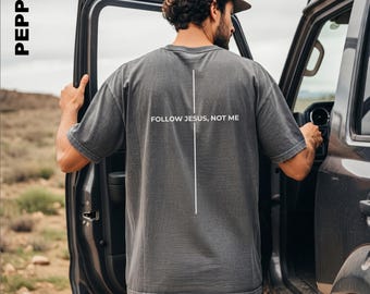 Könnte beinhalten: Ein anthrazitfarbenes Kurzarm-T-Shirt mit einer vertikalen weißen Linie und dem Text "FOLLOW JESUS, NOT ME" auf der Rückseite. Das Shirt hat eine lockere Passform und eine verwaschene Optik, die ein schlichtes Design auf der Rückseite zeigt.