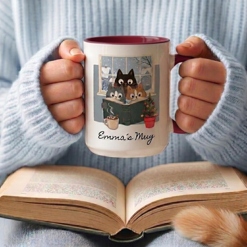 May include: A white ceramic mug with a maroon interior and handle, featuring an illustration of three cats reading a book together, a small mug, and a potted plant. The text "Emma's Mug" is printed in black script below the illustration.