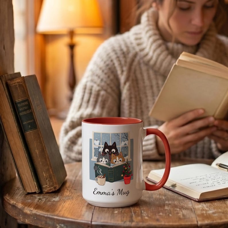May include: A white ceramic mug with a red interior and handle, featuring a graphic of three cats reading a book by a window with snow outside, a small potted tree, and a cup of cocoa. The text "Emma's Mug" is printed in black script below the illustration.
