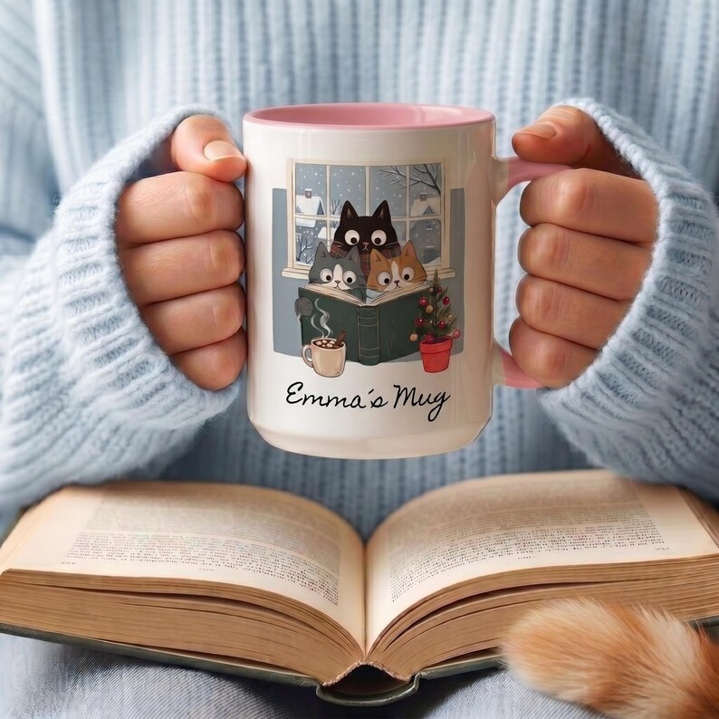 May include: A white ceramic mug with a pink interior and handle, featuring a graphic of three cats reading a book in front of a snowy window, a small mug of cocoa, and a potted plant. The text "Emma's Mug" is printed in black script below the illustration.
