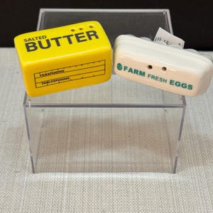 May include: A yellow ceramic butter-shaped salt shaker with the word "BUTTER" in black. A white ceramic egg-shaped salt shaker with the words "FARM FRESH EGGS" in green. Both are displayed in a clear plastic box.
