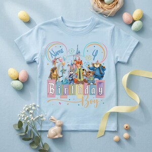 Pu&ograve; includere: Una t-shirt a maniche corte color azzurro con una grafica colorata di personaggi animali animati che indossano cappellini da festa, la sagoma di un castello e il testo Name, 4 e Birthday Boy in un carattere giocoso, centrato sul davanti.