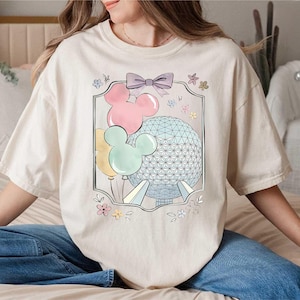 May include: A cream-coloured t-shirt with a framed graphic. The graphic features Mickey Mouse-shaped balloons in pink, yellow, and mint green, a geometric sphere, and a purple bow. The t-shirt has a relaxed fit.