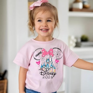 Op de afbeelding: Lichtroze T-shirt met een Disney-kasteelafbeelding en de tekst "Disney 2020". Het ontwerp bevat een roze strik met stippen, muisoren en het woord "NAME" in een cirkel. Een roze strik zit in het haar van het kind.