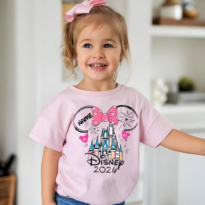 Op de afbeelding: Lichtroze T-shirt met een Disney-kasteelafbeelding en de tekst "Disney 2020". Het ontwerp bevat een roze strik met stippen, muisoren en het woord "NAME" in een cirkel. Een roze strik zit in het haar van het kind.