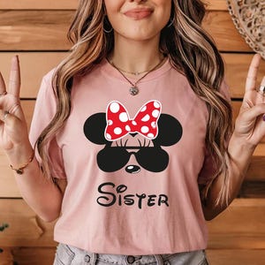 K&ouml;nnte beinhalten: Hellrosa T-Shirt mit Minnie Mouse-Design. Das Design umfasst eine schwarze Sonnenbrille, eine rot-wei&szlig; gepunktete Schleife und das Wort "Sister" in schwarzer Schrift. Das T-Shirt hat einen Rundhalsausschnitt.
