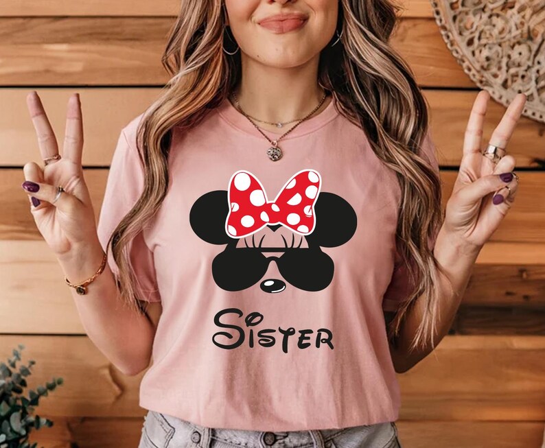 K&ouml;nnte beinhalten: Hellrosa T-Shirt mit Minnie Mouse-Design. Das Design umfasst eine schwarze Sonnenbrille, eine rot-wei&szlig; gepunktete Schleife und das Wort "Sister" in schwarzer Schrift. Das T-Shirt hat einen Rundhalsausschnitt.