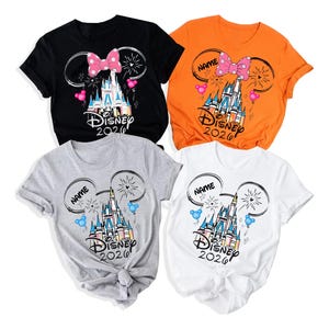 Op de afbeelding: Vier korte mouwen T-shirts in zwart, oranje, grijs en wit. Elk shirt heeft een Disney kasteelafbeelding met een strik, Mickey Mouse oren en de tekst "Disney 2020". De shirts hebben het woord "Name" in een cirkel.