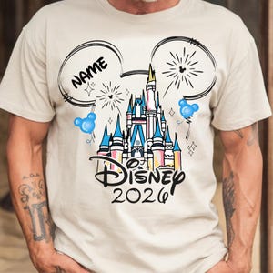 Op de afbeelding: Beige T-shirt met een Disney-kasteelafbeelding in blauw, roze en geel. Het ontwerp bevat Mickey Mouse-oren, vuurwerk en de tekst "Disney 2026" met ruimte voor een naam.