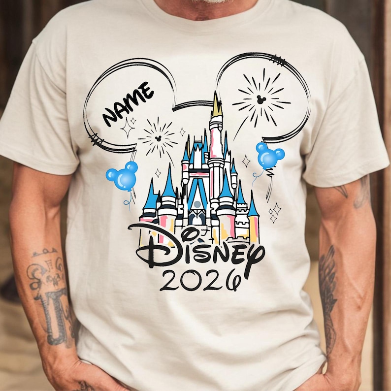 Op de afbeelding: Beige T-shirt met een Disney-kasteelafbeelding in blauw, roze en geel. Het ontwerp bevat Mickey Mouse-oren, vuurwerk en de tekst "Disney 2026" met ruimte voor een naam.