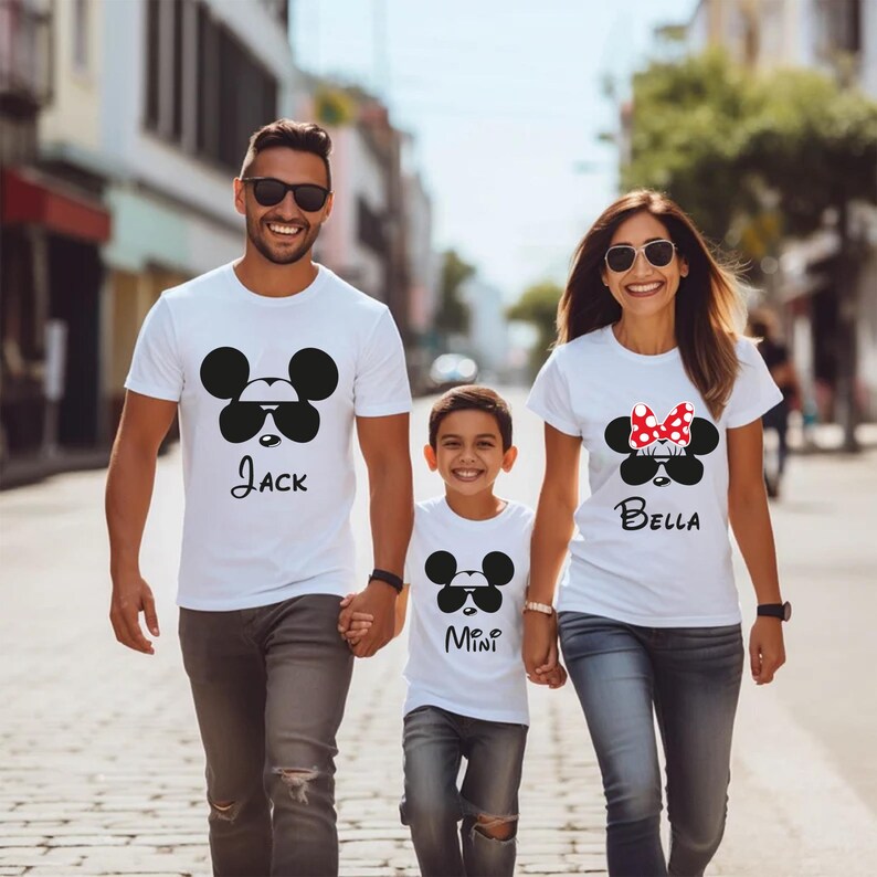 K&ouml;nnte beinhalten: Drei Personen tragen wei&szlig;e T-Shirts mit einem schwarzen Mickey Mouse-Design mit Sonnenbrille. Die Shirts tragen die Namen "Jack", "Mini" und "Bella". Sie gehen eine Stadtstra&szlig;e entlang.