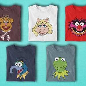 May include: Five folded t-shirts featuring cartoon characters. The shirts are brown, white, and red, with illustrations of Fozzie Bear, Miss Piggy, and Animal. Additional shirts are navy and grey, with Gonzo and Kermit the Frog designs.