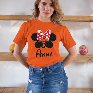 K&ouml;nnte beinhalten: Orangefarbenes T-Shirt mit einem Minnie Mouse-Design, einschlie&szlig;lich einer rot-wei&szlig; gepunkteten Schleife, schwarzer Sonnenbrille und dem Namen "Anna". Das Shirt wird mit einem Jeansrock getragen.