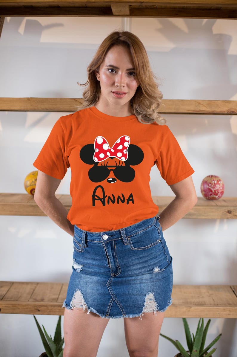K&ouml;nnte beinhalten: Orangefarbenes T-Shirt mit einem Minnie Mouse-Design, einschlie&szlig;lich einer rot-wei&szlig; gepunkteten Schleife, schwarzer Sonnenbrille und dem Namen "Anna". Das Shirt wird mit einem Jeansrock getragen.