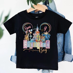 Pu&ograve; includere: Una t-shirt nera a maniche corte con una grafica colorata di personaggi animali animati, un castello e palloncini. Il design include il testo "Name", il numero "4" e le parole "Birthday Boy" in un carattere stilizzato con accenti pastello.
