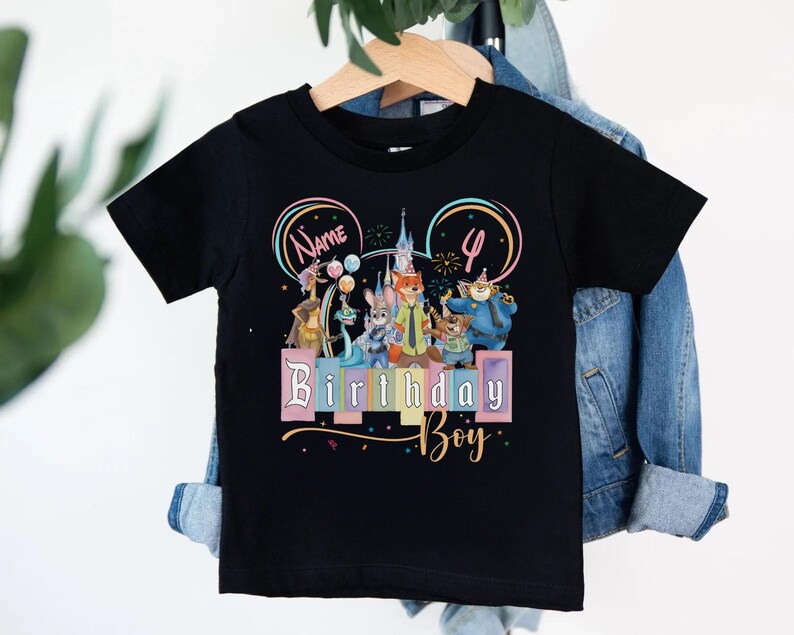 Pu&ograve; includere: Una t-shirt nera a maniche corte con una grafica colorata di personaggi animali animati, un castello e palloncini. Il design include il testo "Name", il numero "4" e le parole "Birthday Boy" in un carattere stilizzato con accenti pastello.