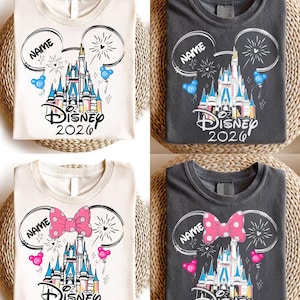 Op de afbeelding: Vier T-shirts met Disney kasteelontwerpen. Twee zijn gebroken wit, twee zijn antracietgrijs. Elk shirt heeft een kasteelafbeelding, de tekst "Disney 2026" en een ruimte voor een naam. Twee shirts hebben Mickey Mouse oren, twee hebben een roze strik met stippen.