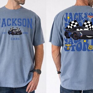 May include: A blue t-shirt featuring a cartoon car design. The front of the shirt displays the text "JACKSON CARS" with a car graphic and the text "EST 2006". The back of the shirt has a similar car graphic with the text "JACKSON STORM" and checkered flags.