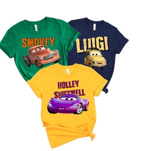 May include: Three short-sleeved t-shirts in green, navy blue, and yellow. Each shirt features a cartoon car graphic and a name: "Smokey", "Luigi", and "Holley Shiftwell". The shirts are made of a soft material.