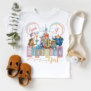 Pu&ograve; includere: Una t-shirt bianca con una grafica colorata di personaggi animali animati, un castello e il testo "Name", "4" e "Birthday Girl". Il design include palloncini, fuochi d'artificio e arcobaleni. Un paio di scarpe marroni &egrave; posizionato accanto.