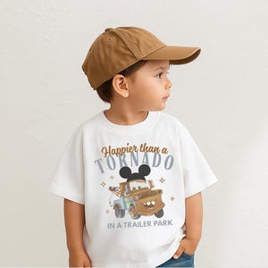 Puede incluir: Un ni&ntilde;o lleva una gorra de b&eacute;isbol marr&oacute;n y una camiseta blanca. La camiseta presenta una imagen de dibujos animados de una gr&uacute;a marr&oacute;n con orejas de Mickey Mouse y el texto "Happier than a Tornado in a Trailer Park."