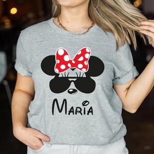 K&ouml;nnte beinhalten: Graues T-Shirt mit einem Minnie-Mouse-Design. Das Design umfasst eine rote Schleife mit wei&szlig;en Punkten, eine schwarze Sonnenbrille und den Namen "Maria" in schwarzer Schrift. Das T-Shirt ist ein l&auml;ssiger Stil.