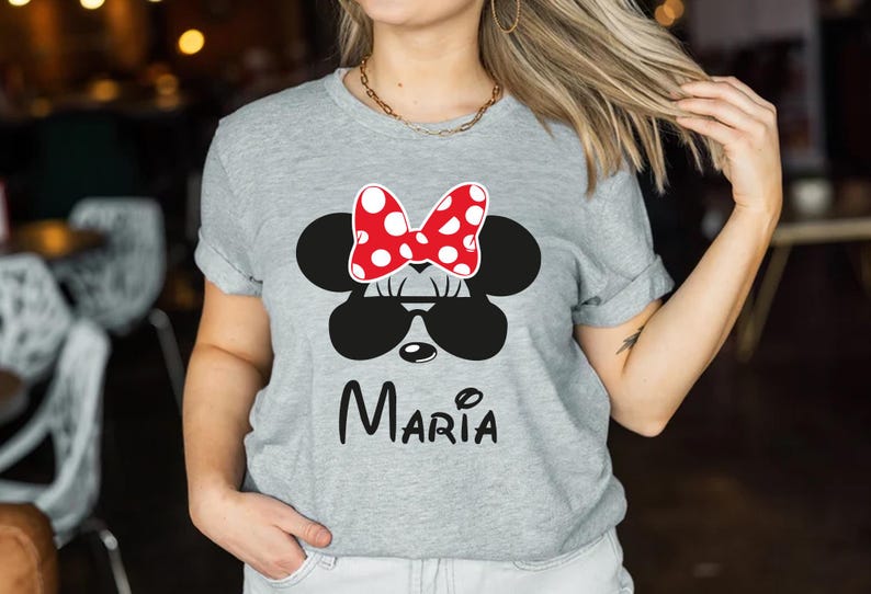 K&ouml;nnte beinhalten: Graues T-Shirt mit einem Minnie-Mouse-Design. Das Design umfasst eine rote Schleife mit wei&szlig;en Punkten, eine schwarze Sonnenbrille und den Namen "Maria" in schwarzer Schrift. Das T-Shirt ist ein l&auml;ssiger Stil.