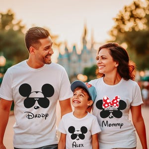 K&ouml;nnte beinhalten: Eine dreik&ouml;pfige Familie tr&auml;gt passende wei&szlig;e T-Shirts mit Mickey- und Minnie-Mouse-Designs. Die Shirts zeigen Sonnenbrillen und die W&ouml;rter "Daddy", "Mommy" und "Alex". Die Familie l&auml;chelt und geht im Freien.