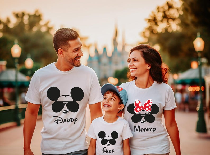 K&ouml;nnte beinhalten: Eine dreik&ouml;pfige Familie tr&auml;gt passende wei&szlig;e T-Shirts mit Mickey- und Minnie-Mouse-Designs. Die Shirts zeigen Sonnenbrillen und die W&ouml;rter "Daddy", "Mommy" und "Alex". Die Familie l&auml;chelt und geht im Freien.