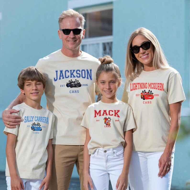 May include: Four people wearing matching cream-colored t-shirts with car-themed designs. The shirts feature names like "Jackson," "Lightning McQueen," "Mack," and "Sally Carrera," along with car graphics and the word "Cars."
