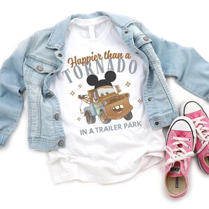 Puede incluir: Camiseta blanca con el texto "Happier than a Tornado in a Trailer Park" y un dise&ntilde;o de cami&oacute;n de remolque de dibujos animados con orejas de Mickey Mouse. La camiseta se combina con una chaqueta vaquera azul claro y zapatillas rosas.