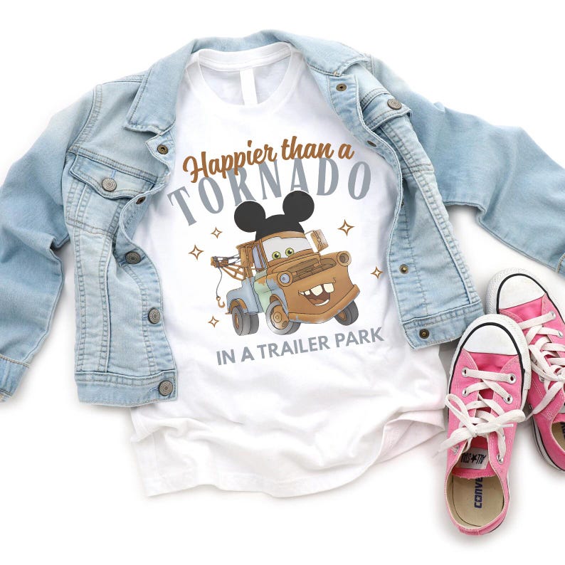 Puede incluir: Camiseta blanca con el texto "Happier than a Tornado in a Trailer Park" y un dise&ntilde;o de cami&oacute;n de remolque de dibujos animados con orejas de Mickey Mouse. La camiseta se combina con una chaqueta vaquera azul claro y zapatillas rosas.