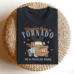 Puede incluir: Camiseta negra con un dise&ntilde;o de cami&oacute;n de remolque de dibujos animados y el texto "Happier than a Tornado in a Trailer Park". El dise&ntilde;o incluye estrellas y una silueta de Mickey Mouse. La camiseta se muestra sobre una superficie tejida.