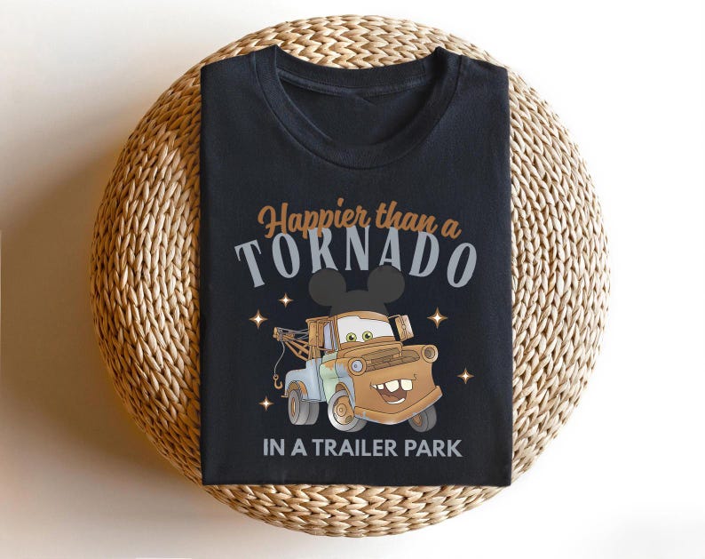 Puede incluir: Camiseta negra con un dise&ntilde;o de cami&oacute;n de remolque de dibujos animados y el texto "Happier than a Tornado in a Trailer Park". El dise&ntilde;o incluye estrellas y una silueta de Mickey Mouse. La camiseta se muestra sobre una superficie tejida.