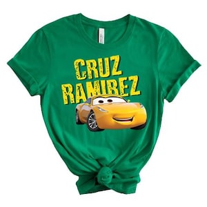 May include: A green t-shirt featuring the text "CRUZ RAMIREZ" in yellow, with a yellow cartoon car graphic underneath. The t-shirt is made of a soft material and has short sleeves.