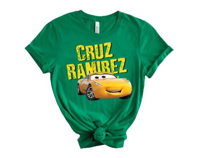May include: A green t-shirt featuring the text "CRUZ RAMIREZ" in yellow, with a yellow cartoon car graphic underneath. The t-shirt is made of a soft material and has short sleeves.