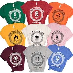 May include: A collection of colorful t-shirts featuring the text "Camp Half-Blood" in a circular design. Each shirt has a different color and a symbol representing a Greek god or goddess, such as Zeus, Ares, and Athena. The shirts are short-sleeved.