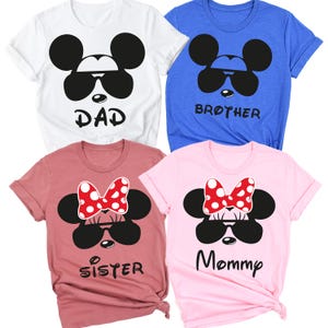 K&ouml;nnte beinhalten: Vier kurz&auml;rmelige T-Shirts in Wei&szlig;, Blau, Mauve und Rosa. Jedes Shirt hat ein schwarzes, von Mickey Mouse inspiriertes Design mit Sonnenbrille. Das wei&szlig;e Shirt sagt "DAD", das blaue Shirt sagt "BROTHER", das mauvefarbene Shirt sagt "SISTER" und das rosa Shirt sagt "Mommy".