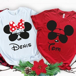 K&ouml;nnte beinhalten: Zwei kurz&auml;rmelige T-Shirts, eines wei&szlig; und eines rot, mit Cartoon-Mausgesichtern mit Sonnenbrillen. Das wei&szlig;e Shirt hat eine rote Schleife mit Punkten und den Namen "Denis". Das rote Shirt hat den Namen "Tom".