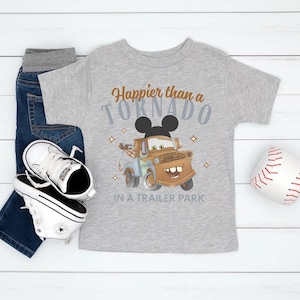 Puede incluir: Una camiseta gris con el texto "Happier than a Tornado in a Trailer Park" y un dise&ntilde;o de cami&oacute;n de remolque de dibujos animados. La imagen tambi&eacute;n muestra vaqueros azules, zapatillas blancas y una pelota de b&eacute;isbol.