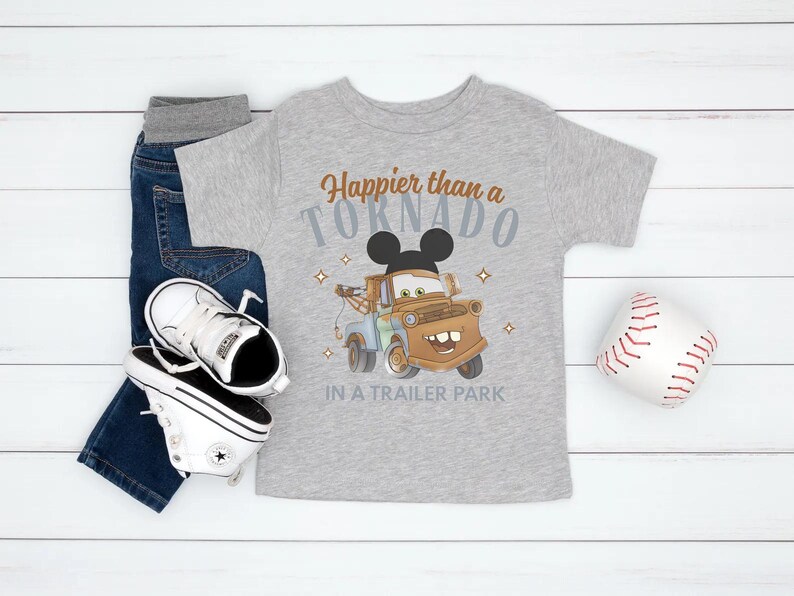 Puede incluir: Una camiseta gris con el texto "Happier than a Tornado in a Trailer Park" y un dise&ntilde;o de cami&oacute;n de remolque de dibujos animados. La imagen tambi&eacute;n muestra vaqueros azules, zapatillas blancas y una pelota de b&eacute;isbol.